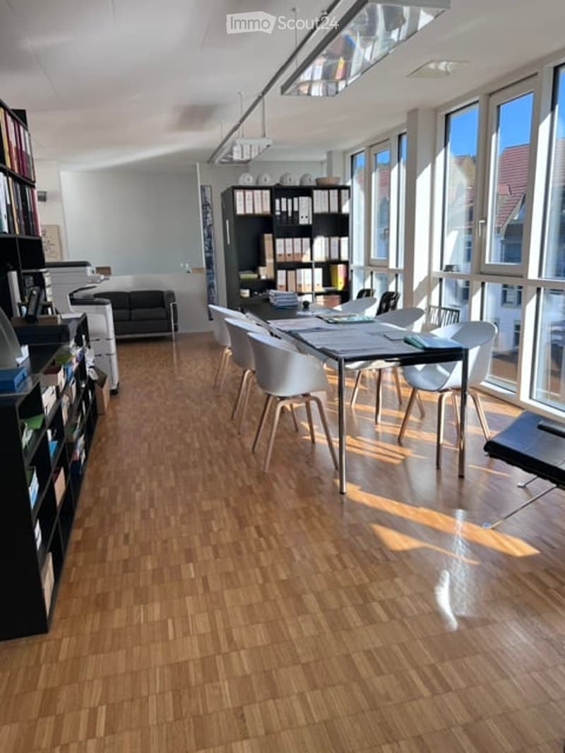 Bright, modern office or meeting space with wooden flooring, large windows, a long conference table with chairs, shelves with books and office supplies, and a black sofa in the background.