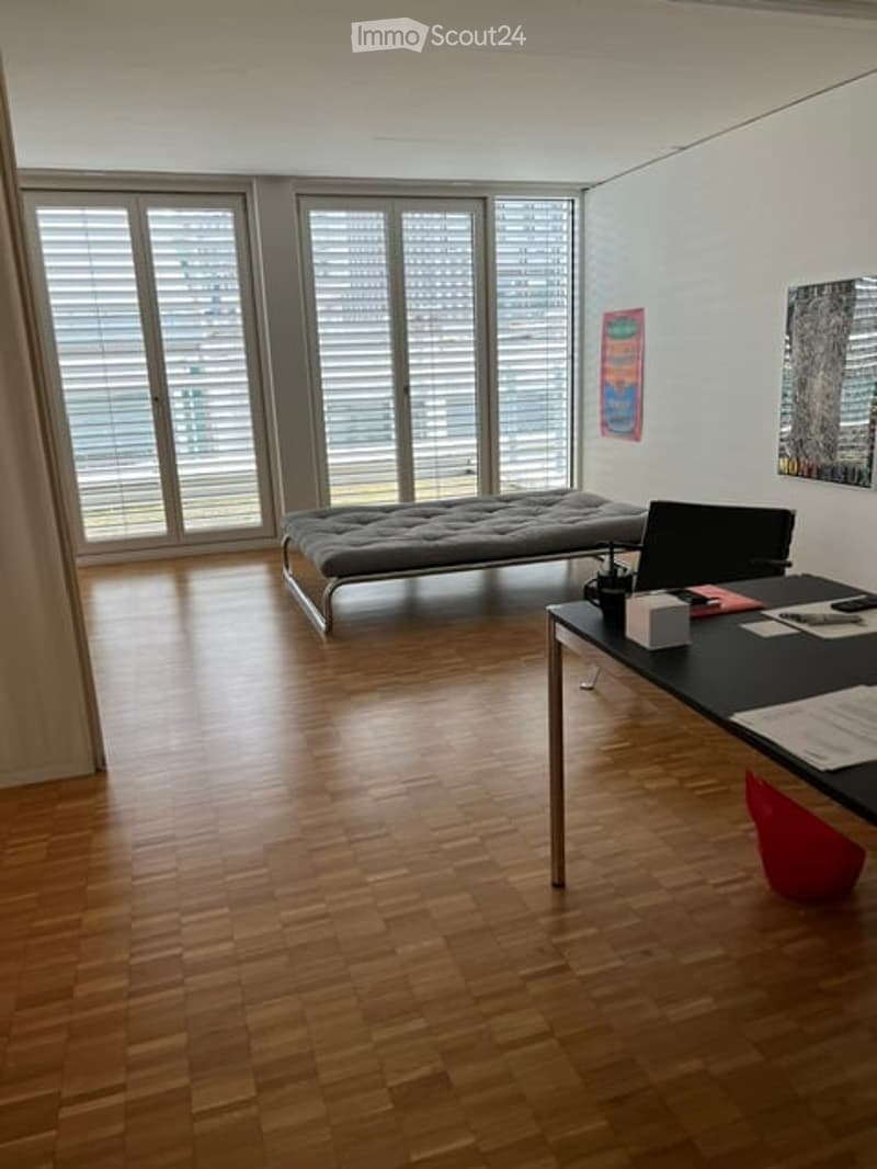 Modern, minimalist room with wooden flooring, large windows with blinds, a sofa, a desk with a laptop, and a red chair.