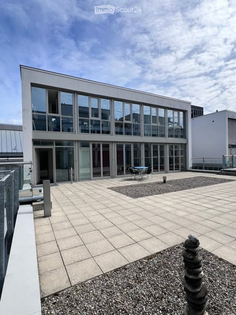 Modern building with large glass windows and a spacious outdoor terrace area featuring a small seating area with a table and chairs.