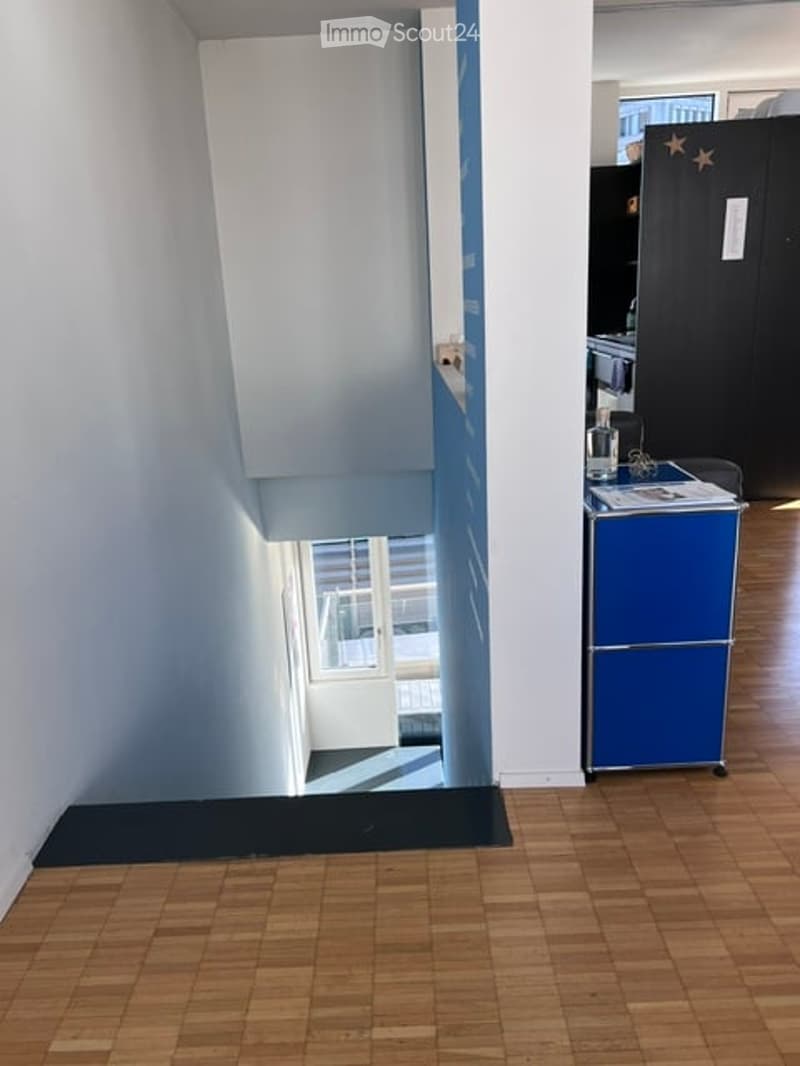 A narrow hallway with wooden flooring, a small blue refrigerator, a stool, and a small table with papers and a glass. The hallway leads to a room with white walls and a window.