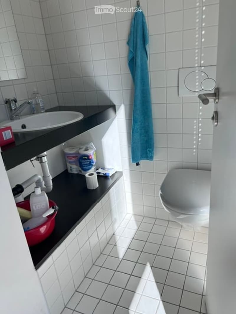Small bathroom with white tiled walls and floor, black countertop, sink, faucet, toilet, towel, and various toiletries.