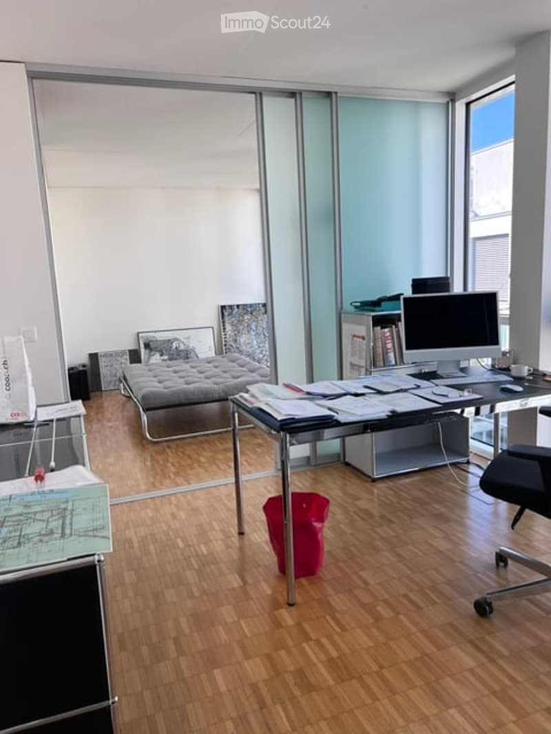 A modern, well-lit room with a wooden floor, large windows, a desk with a computer setup, a bed, and several artworks on the walls.