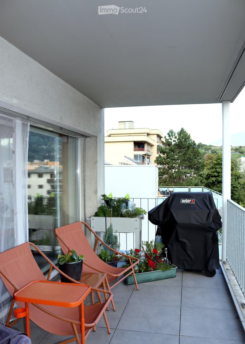 Balcony with two orange chairs, a small table, potted plants, and a covered grill, overlooking a residential area with trees and buildings.