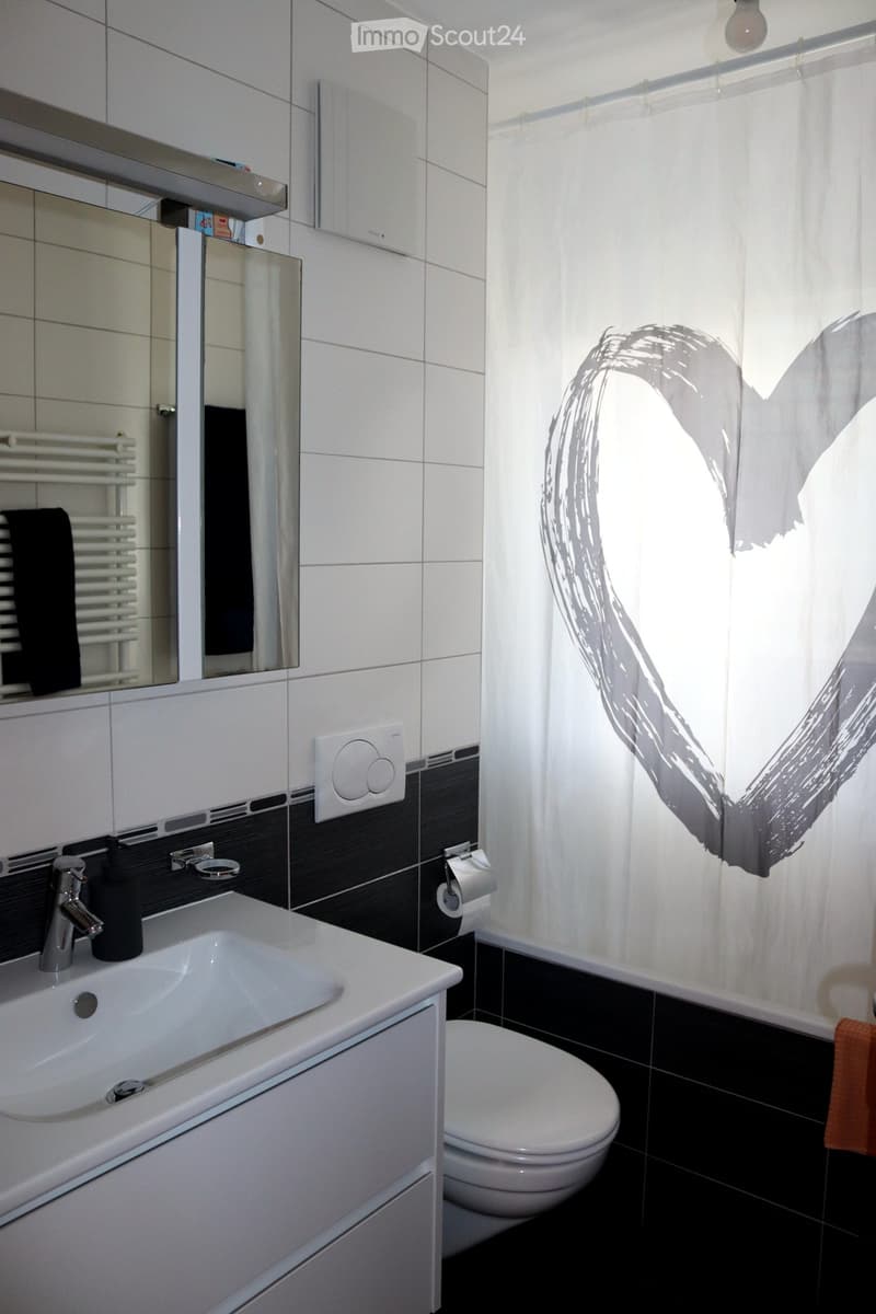 Modern bathroom with white tiles, black accents, a sink with a chrome faucet, a toilet, a mirror, a radiator, and a towel visible.