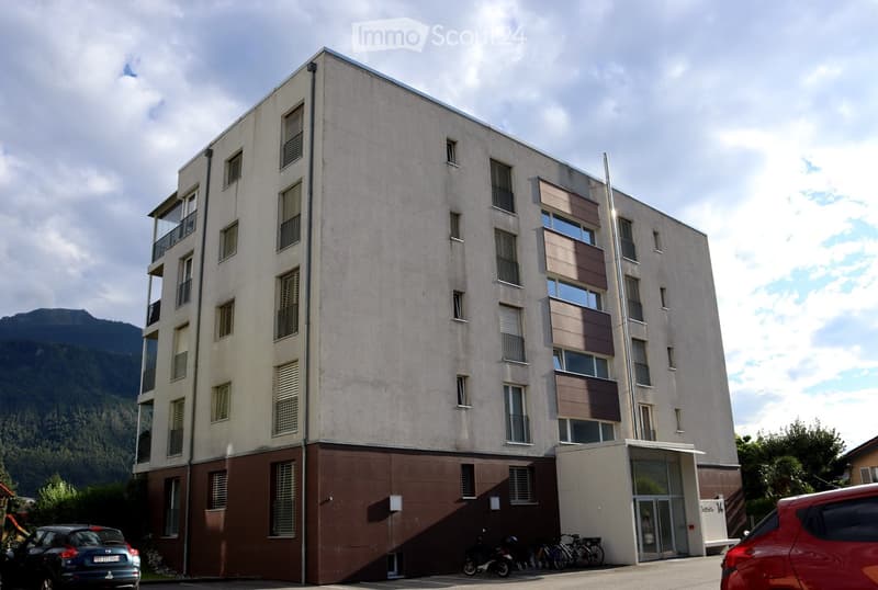 Modern multi-story building with beige and brown facade, multiple windows, balconies, and a parking area with several bicycles and a scooter.