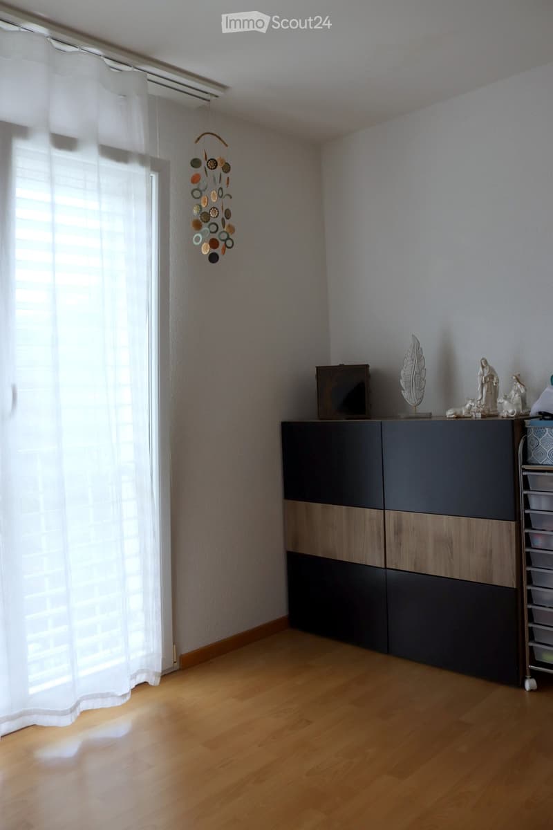 A modern, minimally furnished room with light wooden flooring, a large window with white curtains, a dark wood and grey cabinet, a decorative wind chime, and a small sculpture on top of the cabinet.