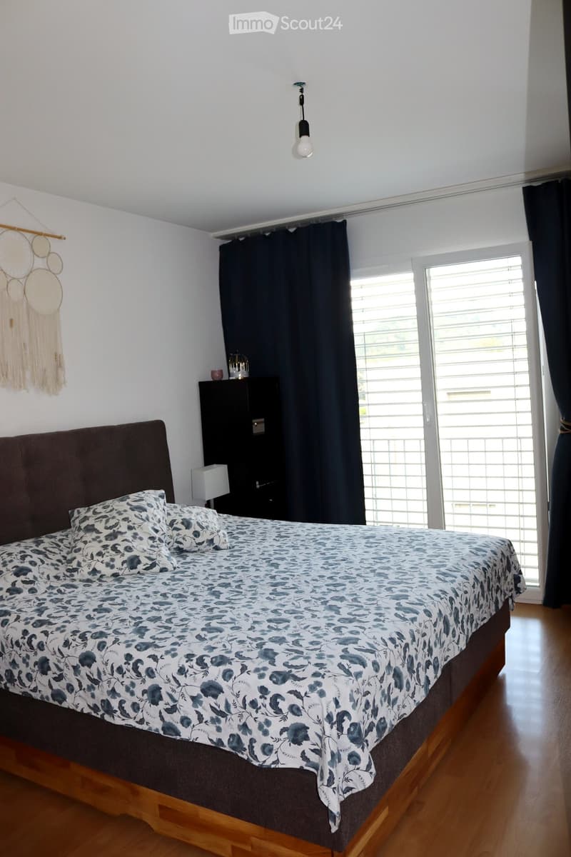 A well-lit bedroom with a large bed covered in a blue and white floral duvet, a nightstand with a lamp, and a window with dark curtains. A dreamcatcher hangs on the wall.