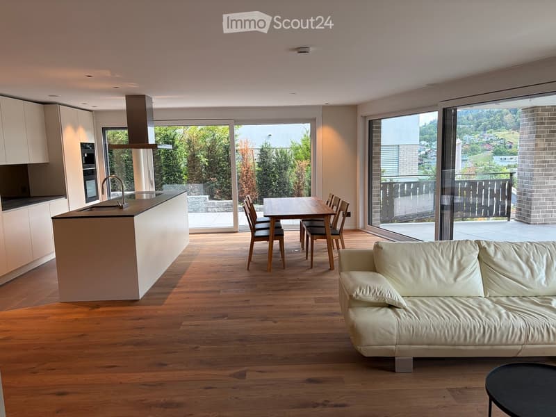 Open-plan living and dining area with a cream sofa, coffee table, dining table with chairs, and a modern kitchen island with an oven and faucet. Large windows offer a scenic view of greenery and a balcony.
