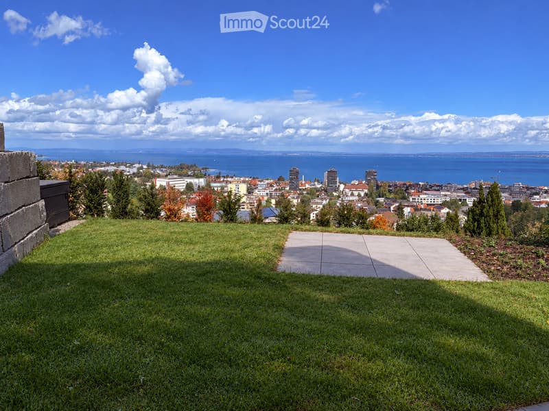 A grassy area with a paved pathway leading towards a cityscape view, with trees and buildings visible in the distance under a partly cloudy sky.