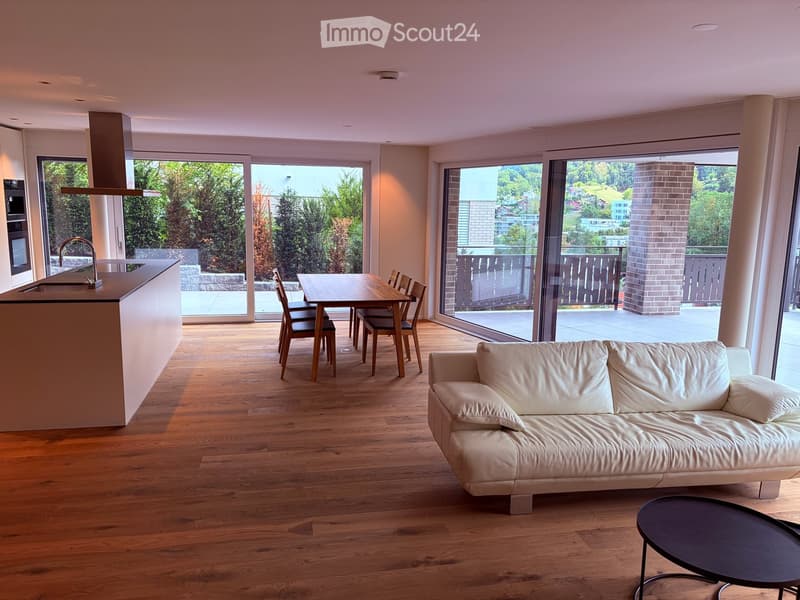 Open-plan living and dining area with a white leather sofa, coffee table, dining table with chairs, and a kitchen island with a faucet. Large windows offer a scenic view of greenery and a balcony.
