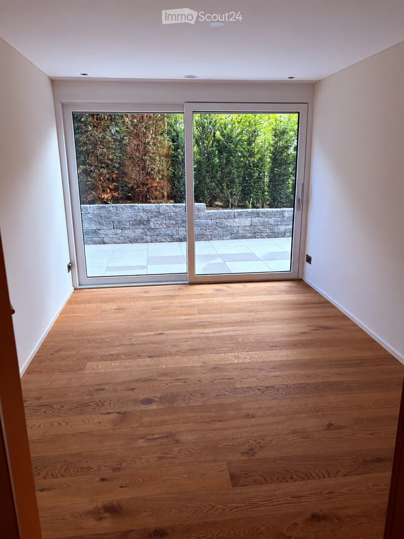 Empty room with wooden flooring, white walls, and large sliding glass doors leading to an outdoor area with a stone wall and greenery.