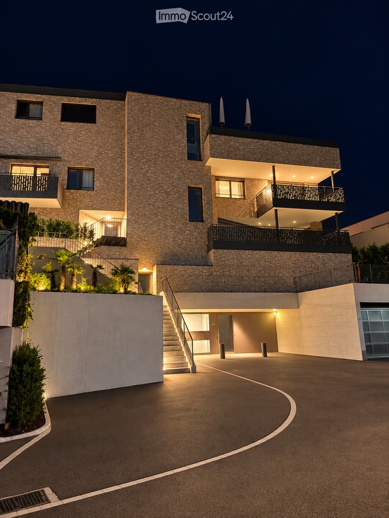 Modern two-story house with brick facade, illuminated windows, balconies, and a curved driveway leading to a garage entrance.