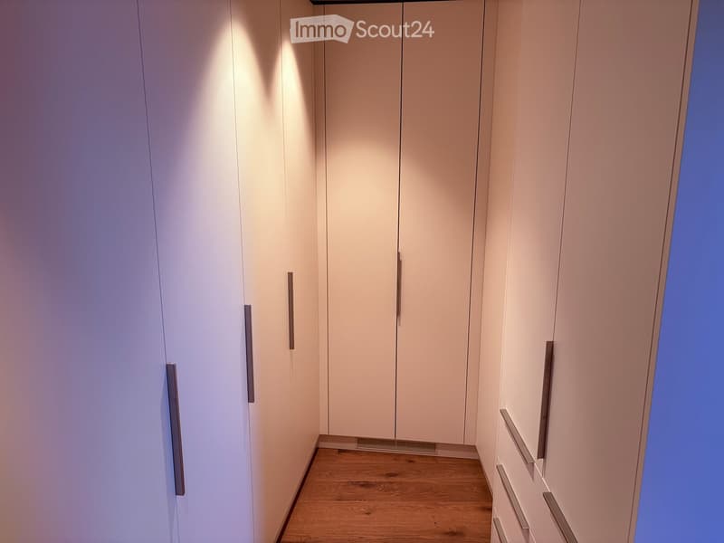 A narrow hallway with light-colored, modern built-in wardrobes and a wooden floor. The wardrobes have sleek, metallic handles and are illuminated by soft, ambient lighting.