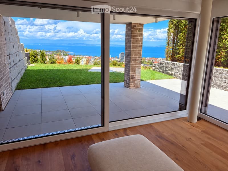 Modern interior with wooden flooring, large glass sliding doors opening to a balcony with scenic ocean view, and a light-colored sofa visible.