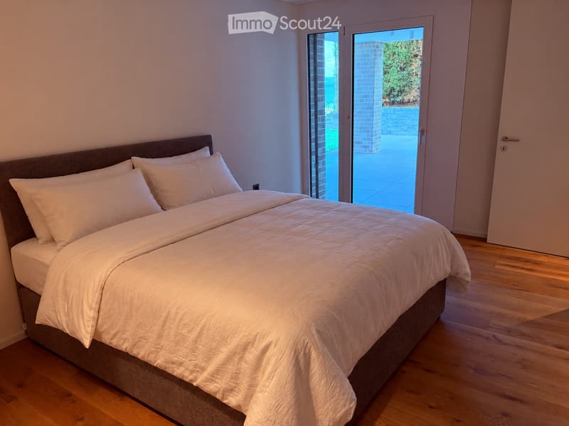 Modern bedroom with a large bed, white bedding, and wooden flooring. Large sliding glass doors lead to an outdoor area.
