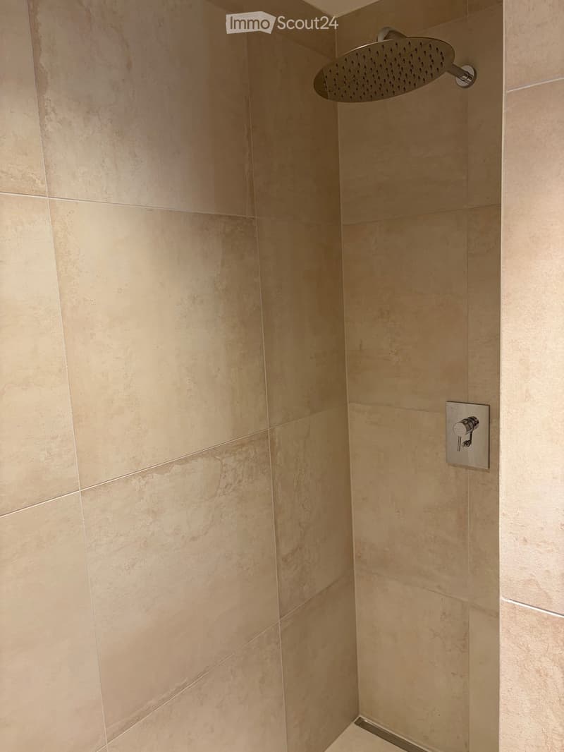 Modern bathroom corner with beige tiled walls, a round showerhead mounted on the wall, and a chrome control valve.