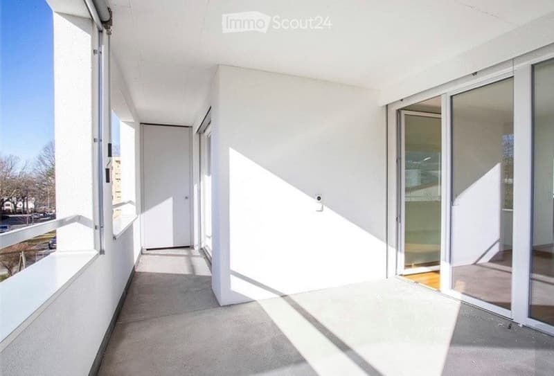 Modern balcony with white walls, large glass sliding doors, and a smooth concrete floor. Sunlight streams in, creating bright shadows on the surface.