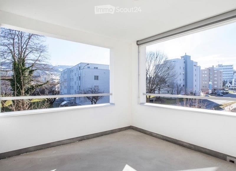 Empty room with large windows offering a view of the city and surrounding buildings. The space has white walls and a concrete floor, with no furniture or decorations present.