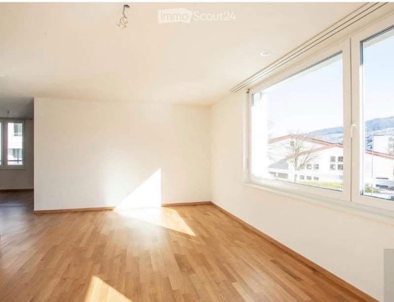 Empty room with wooden flooring and large windows allowing natural light to enter. The walls are painted white, and there are no furnishings or decorations visible.