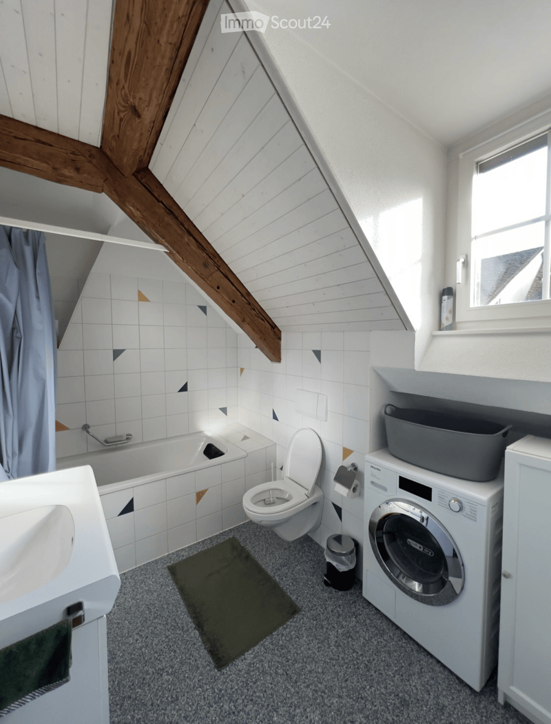 Cozy attic bathroom with white tiled walls, a bathtub, toilet, sink, washing machine, and a small window letting in natural light.
