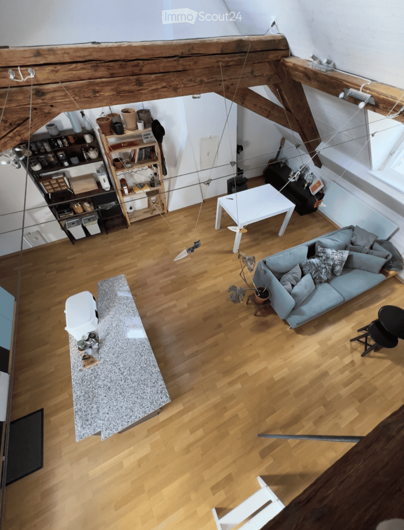 A modern, open-concept living space with wooden flooring, a light blue sofa, a white coffee table, a small white table, a shelf with books and decorative items, and a small kitchen area with a countertop.