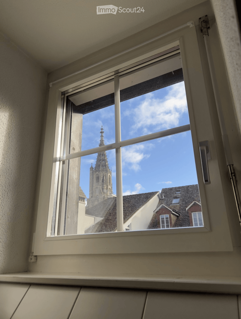 Empty room with a large window showing a view of a church tower and surrounding rooftops.