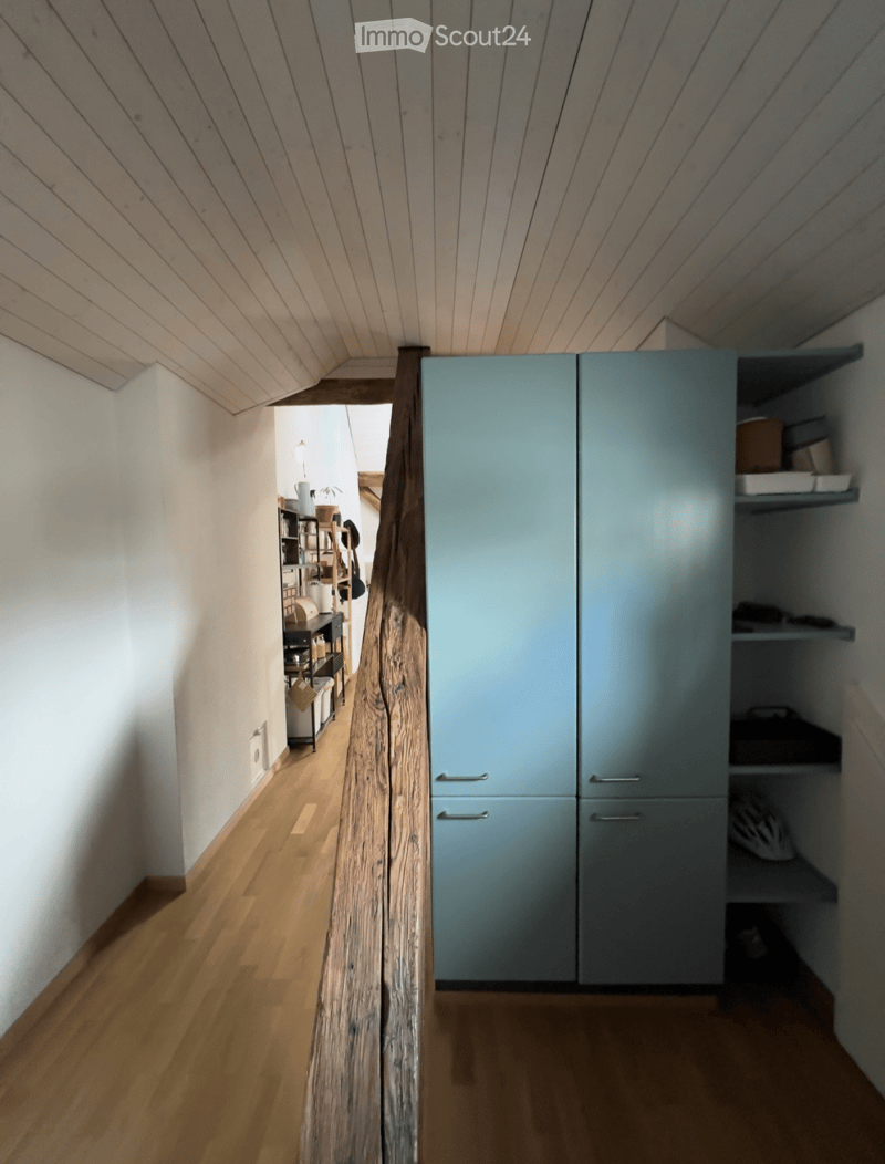 A narrow hallway with wooden flooring, white walls, and a slanted wooden ceiling. A light blue cabinet with shelves is visible on the right side, and a small shelf with various items is seen in the background on the left side.
