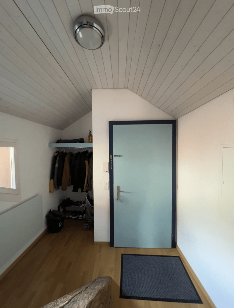 A narrow hallway with wooden flooring, white walls, a slanted ceiling, a round ceiling light fixture, a light blue door, a small shelf with hanging coats, and a dark blue doormat.