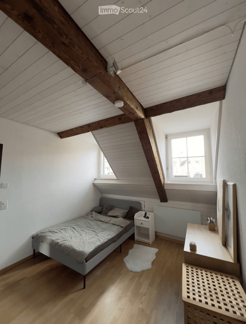 Cozy attic bedroom with wooden slanted ceilings, a bed with pillows and blankets, a nightstand, a desk with a chair, and a small rug on the floor.