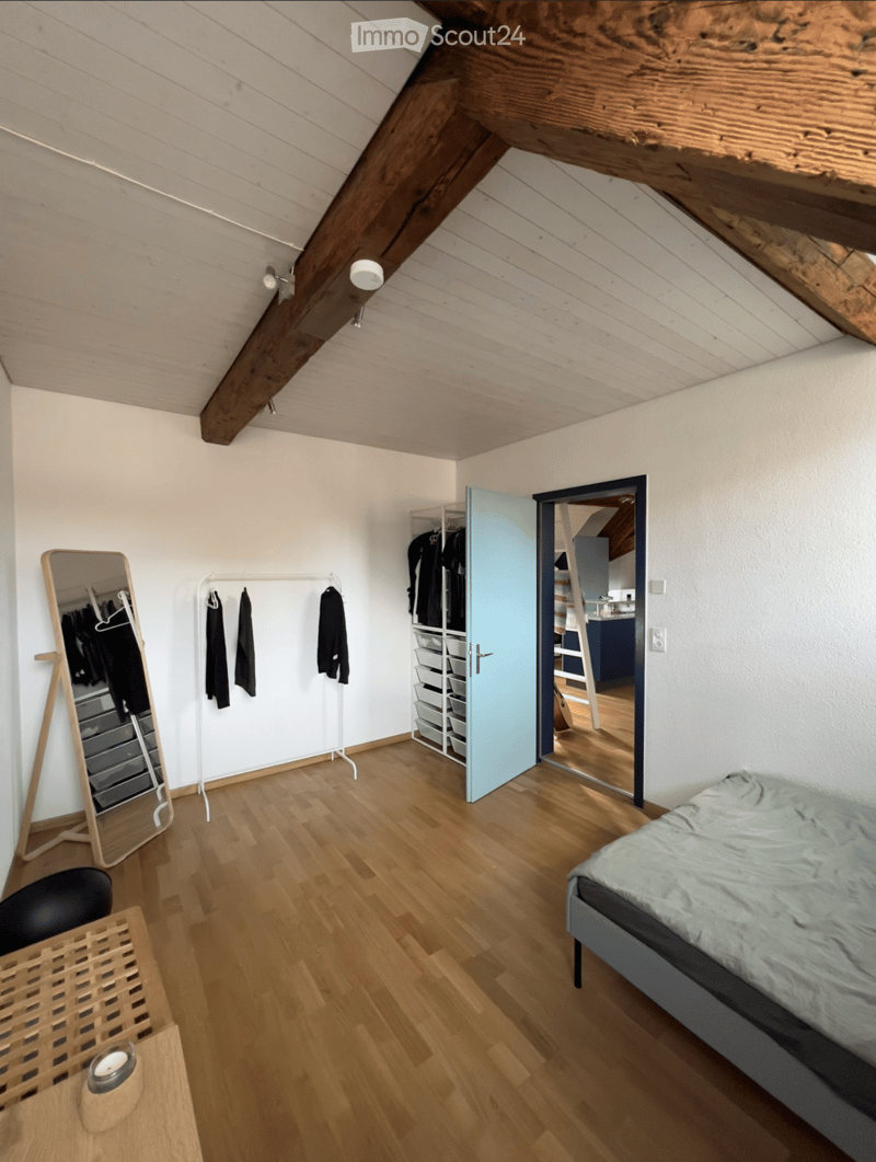 Cozy attic bedroom with wooden floors, slanted wooden ceiling beams, a bed with grey bedding, a full-length mirror, a clothing rack, and a small storage unit.