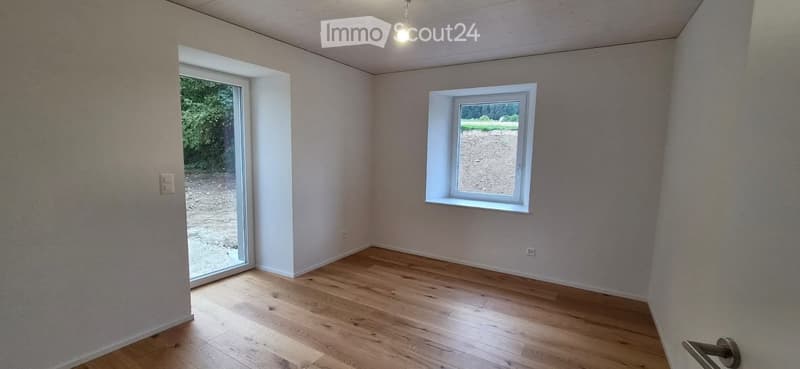 Empty room with wooden flooring, white walls, and two windows, one of which is a sliding glass door leading outside.