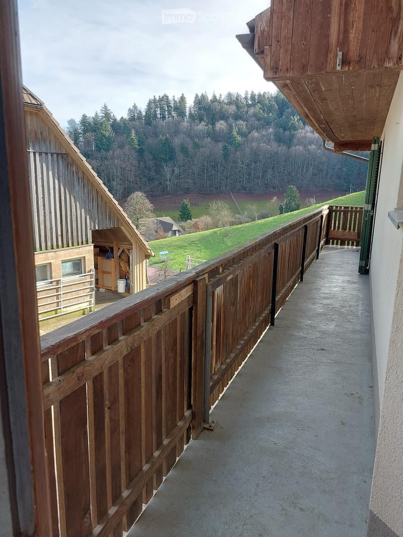 Balcony with wooden railing, overlooking a green landscape with trees and a small building in the distance.