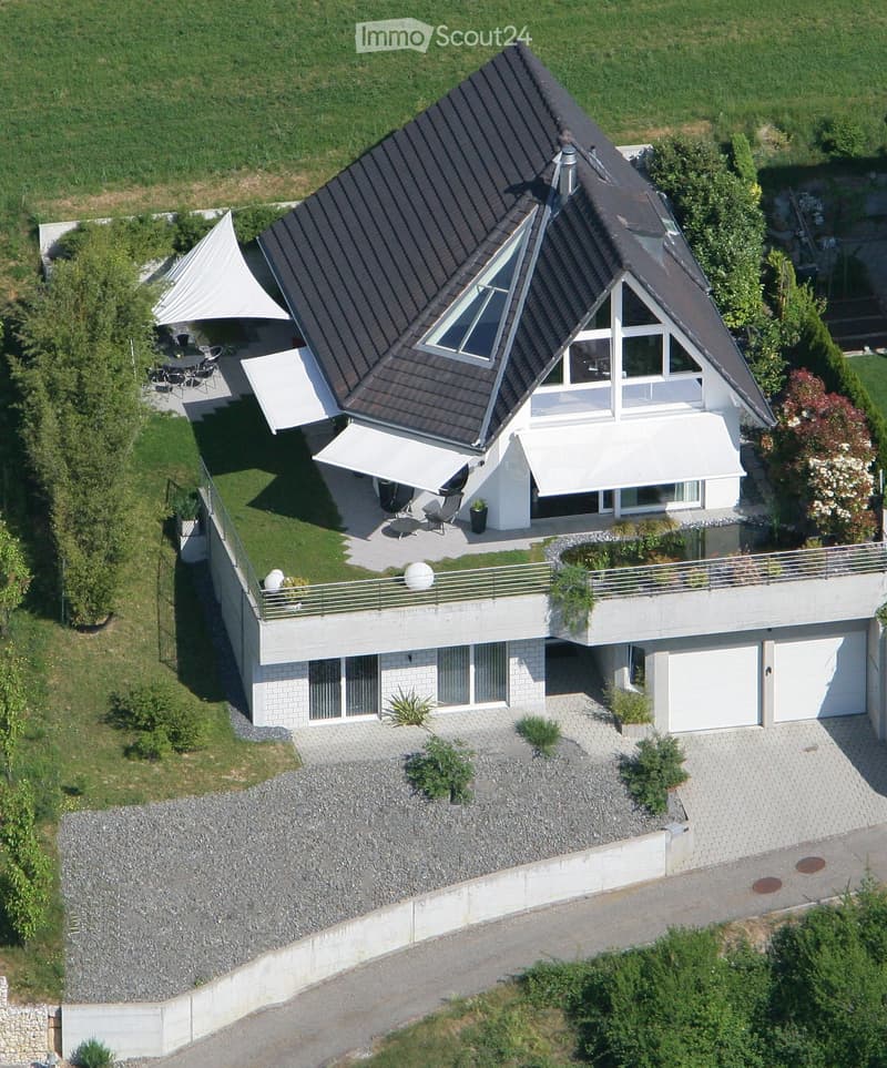 Two-story house with a modern design, featuring a dark roof, large windows, a balcony, and a garage. The surrounding area includes a well-maintained lawn, patio furniture, and a raised ground floor.