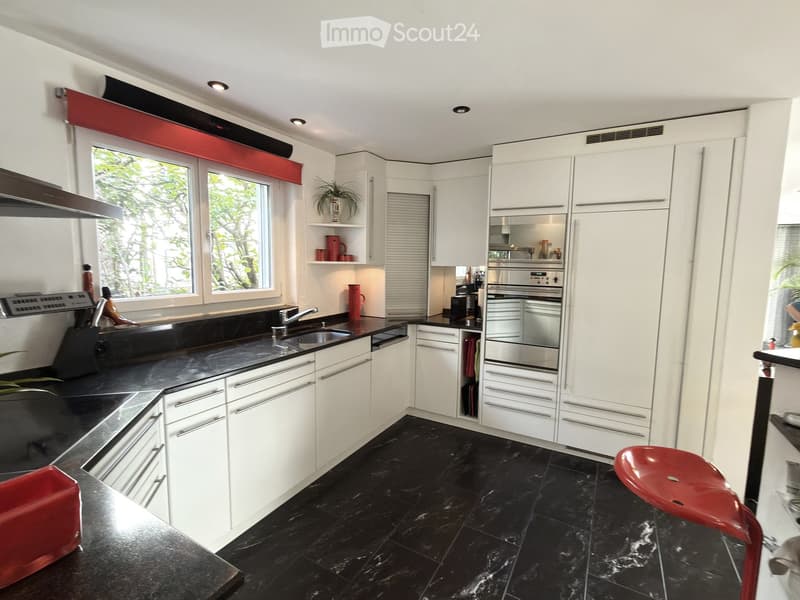 Modern kitchen with white cabinets, black countertops, and black tiled floor. Features a sink, faucet, oven, and a red stool.