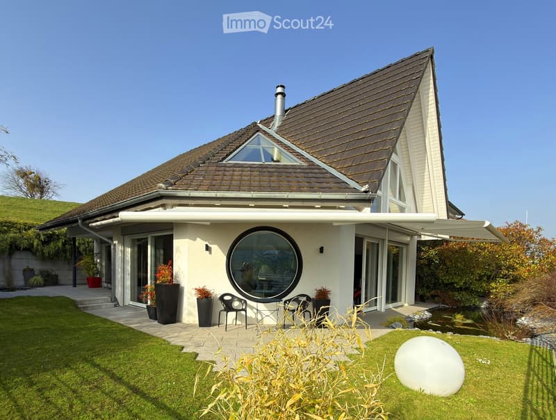 Modern two-story house with a brown tiled roof, large windows, and a spacious garden featuring a pond, patio seating, and decorative plants.