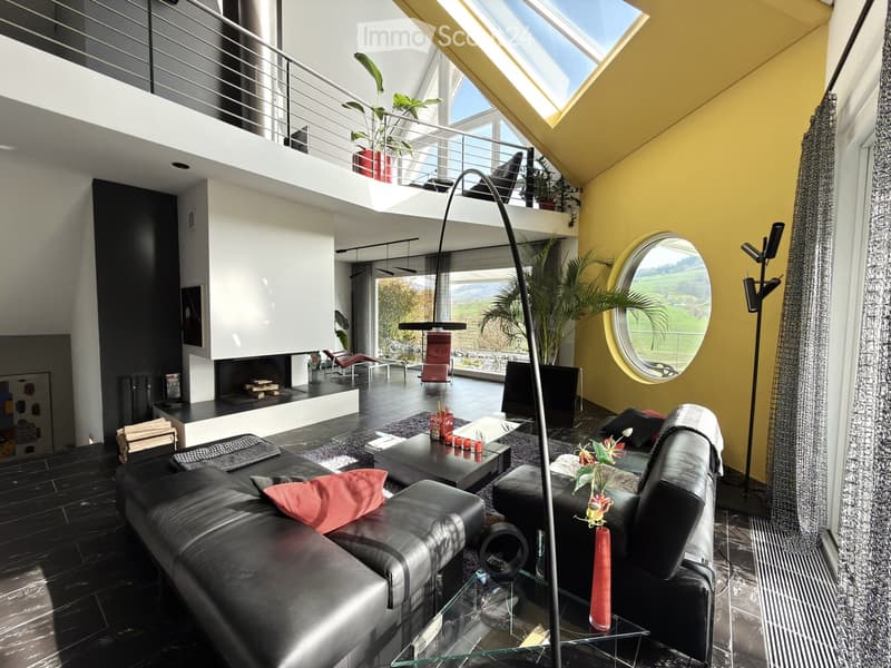 Modern living room with black leather sofa, coffee table, fireplace, large windows offering a scenic view, and decorative elements including a floor lamp and a mirror.