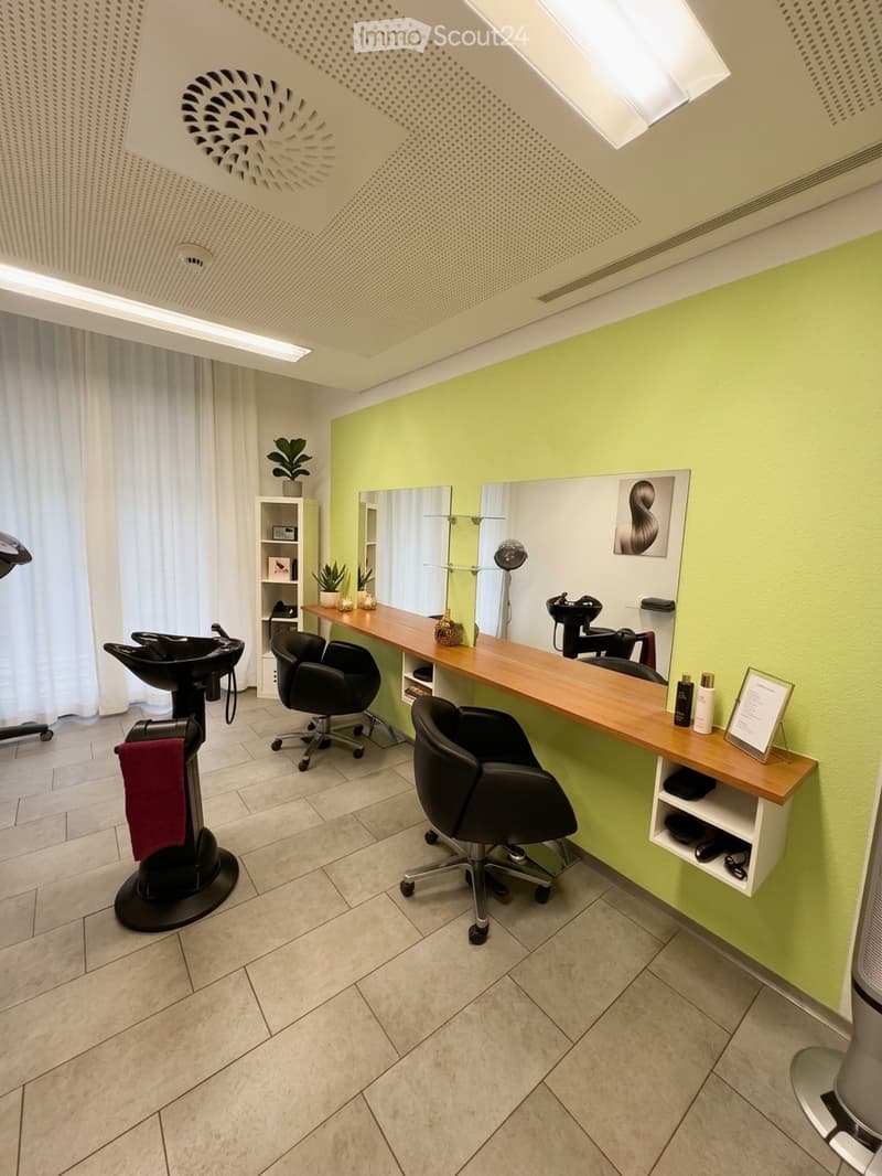 Modern, well-lit salon with light green walls, wooden counter, black chairs, mirrors, shelves, and decorative plants.