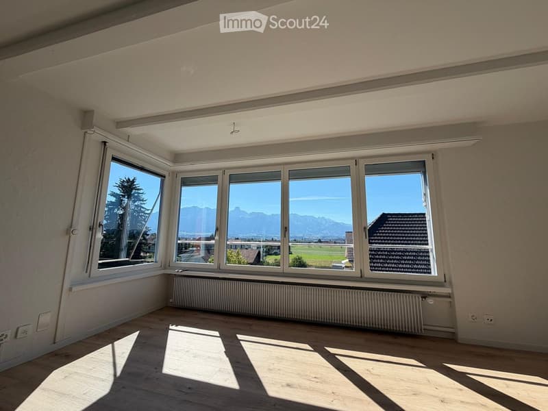 Bright, empty room with wooden flooring, large windows offering a scenic mountain view, and a radiator below the windows.