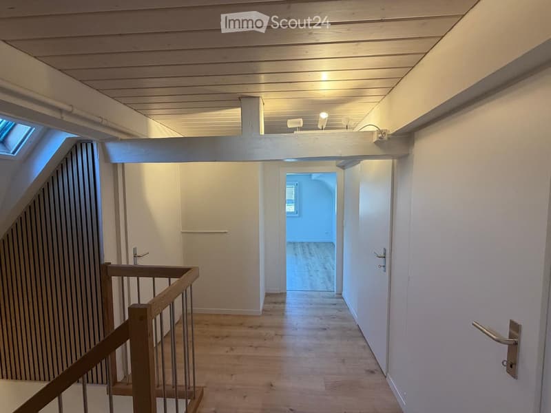 Bright hallway with wooden flooring, white walls, and a staircase leading upwards. Features include recessed ceiling lighting, a skylight, and doors leading to other rooms.