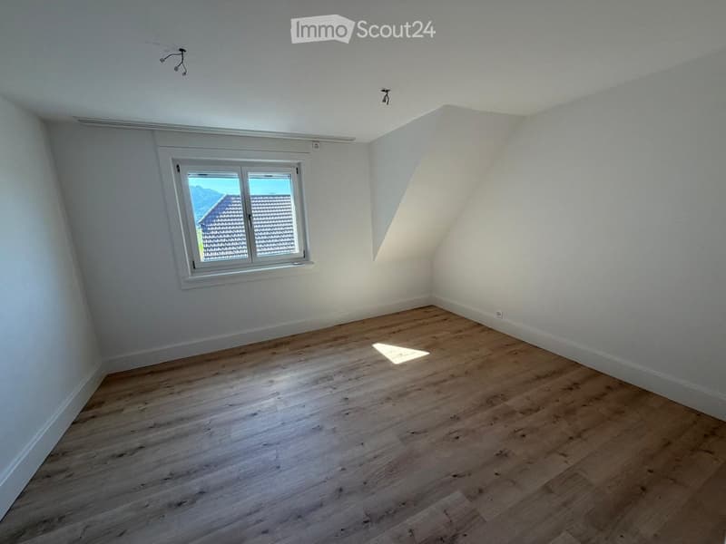 Empty attic room with wooden flooring, white walls, and a single window showing a view of a house and mountains.