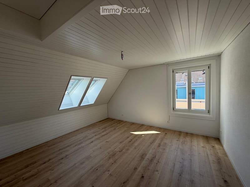 Empty attic room with wooden flooring, white paneled walls, and multiple windows including a skylight and a large window with a view of the street.