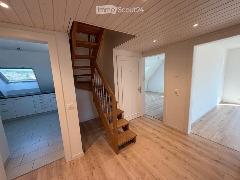 Bright hallway with wooden flooring, a staircase leading upwards, and an open doorway to another room. The walls are painted in a light neutral color, and the ceiling has recessed lighting.