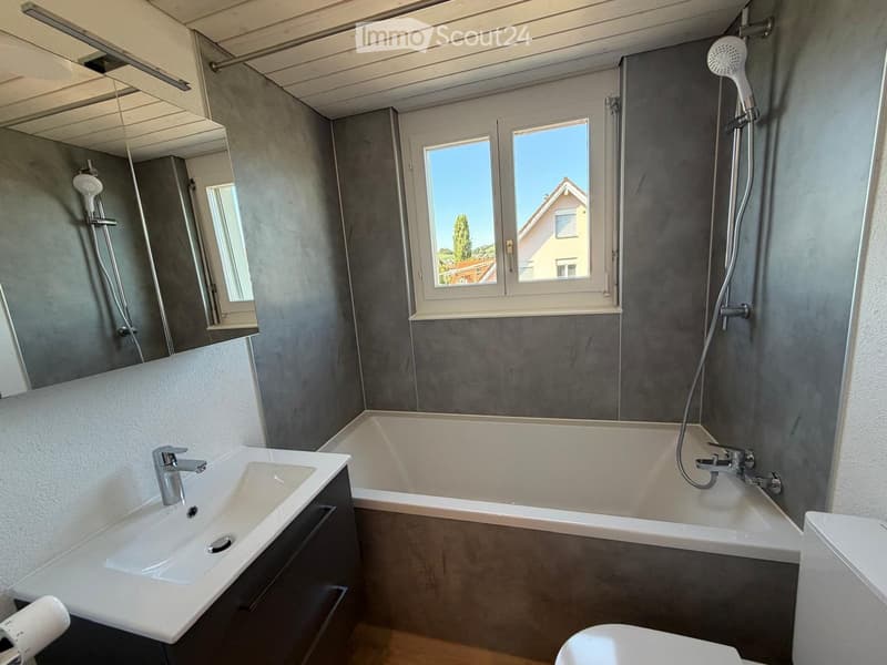 Modern bathroom with a white sink, bathtub, toilet, and showerhead. Grey walls and large windows provide natural light.