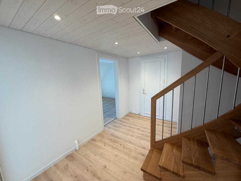 Modern hallway with wooden flooring, white walls, recessed ceiling lights, and a wooden staircase leading upwards.