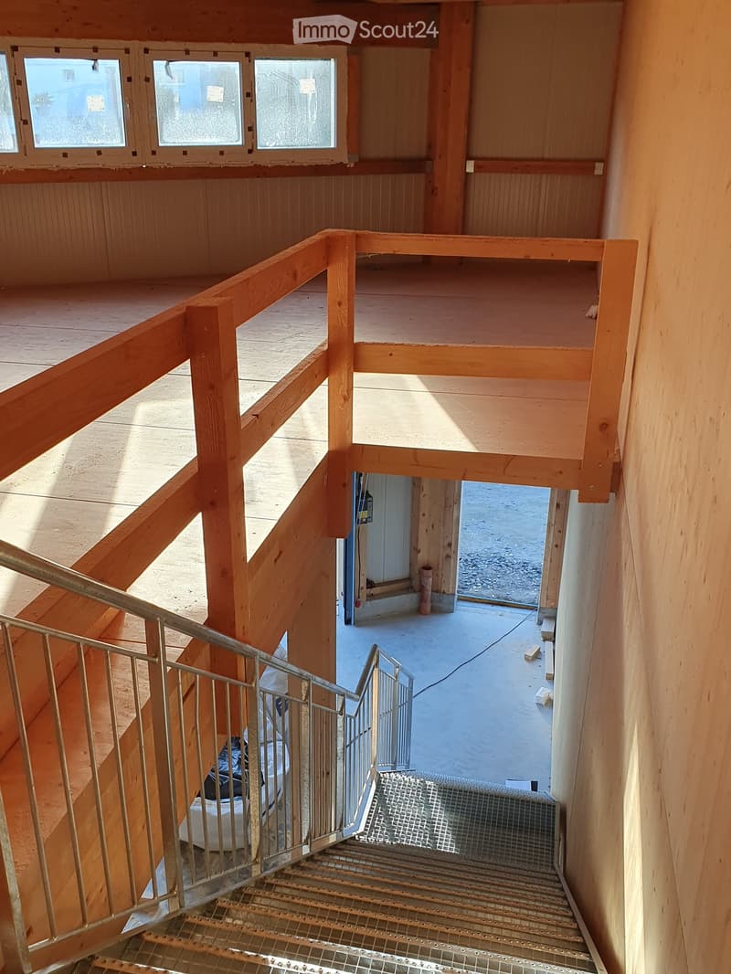 Wooden staircase with metal railing, leading to an open area with wooden walls and windows, showing a partially constructed interior space.