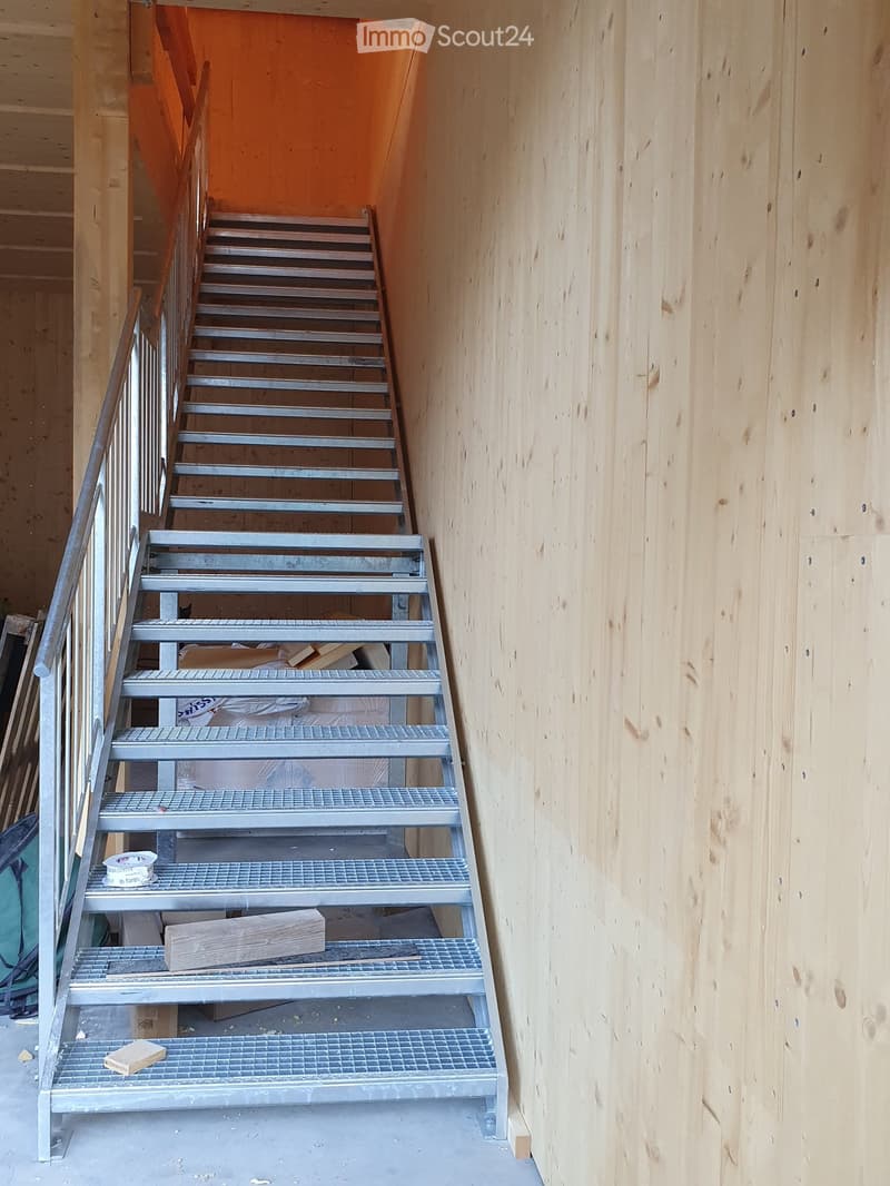 A narrow, unfinished staircase with metal steps and a wooden handrail, leading upwards. The walls are unfinished wooden panels, and the floor is concrete.