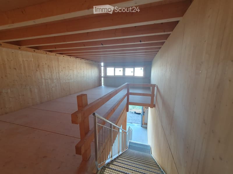 Empty wooden hallway with exposed beams, a metal staircase, and a wooden balcony overlooking an open space with windows.