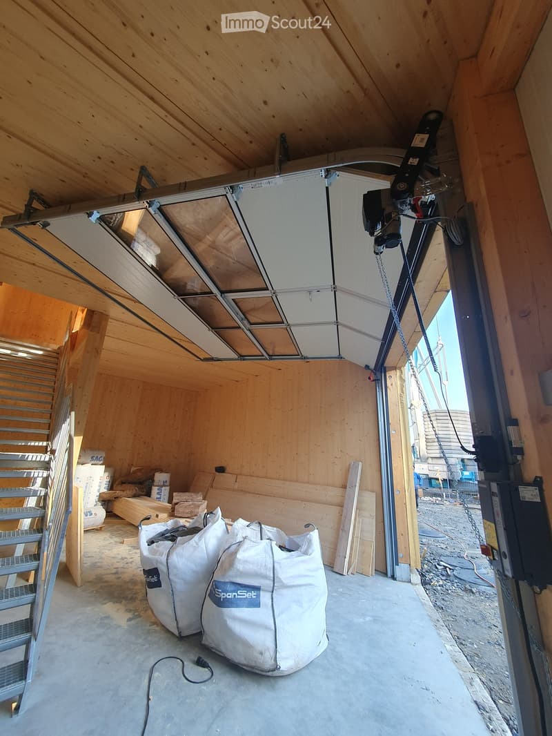 Empty garage with wooden walls, concrete floor, and a partially open garage door. There are large white bags, wooden planks, and a metal ladder visible.