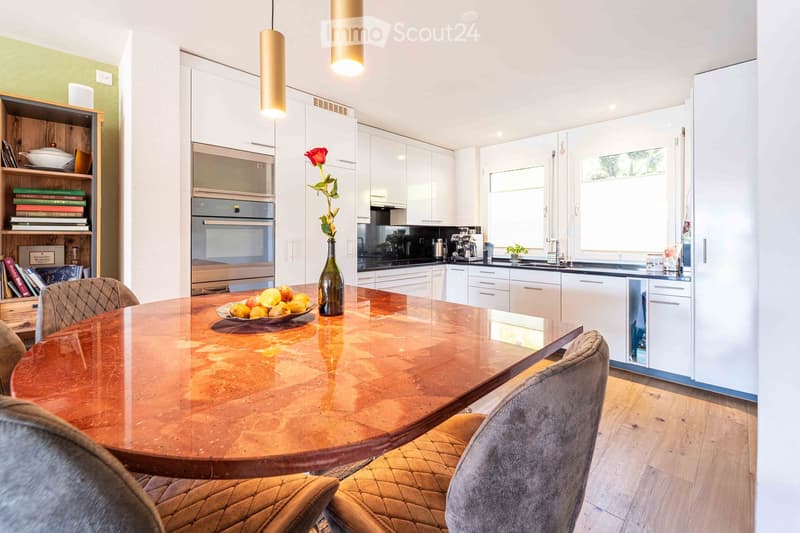 Modern kitchen with white cabinets, a round wooden dining table, and a vase with a red rose on the table. The kitchen features an oven, a coffee machine, and a bookshelf with books and decorative items.