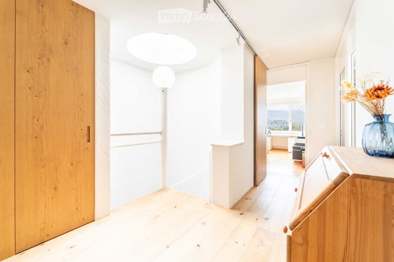 Bright, modern hallway with light wooden flooring, white walls, and a round skylight. Features a wooden door on the left and a vase with dried flowers on a console table to the right.